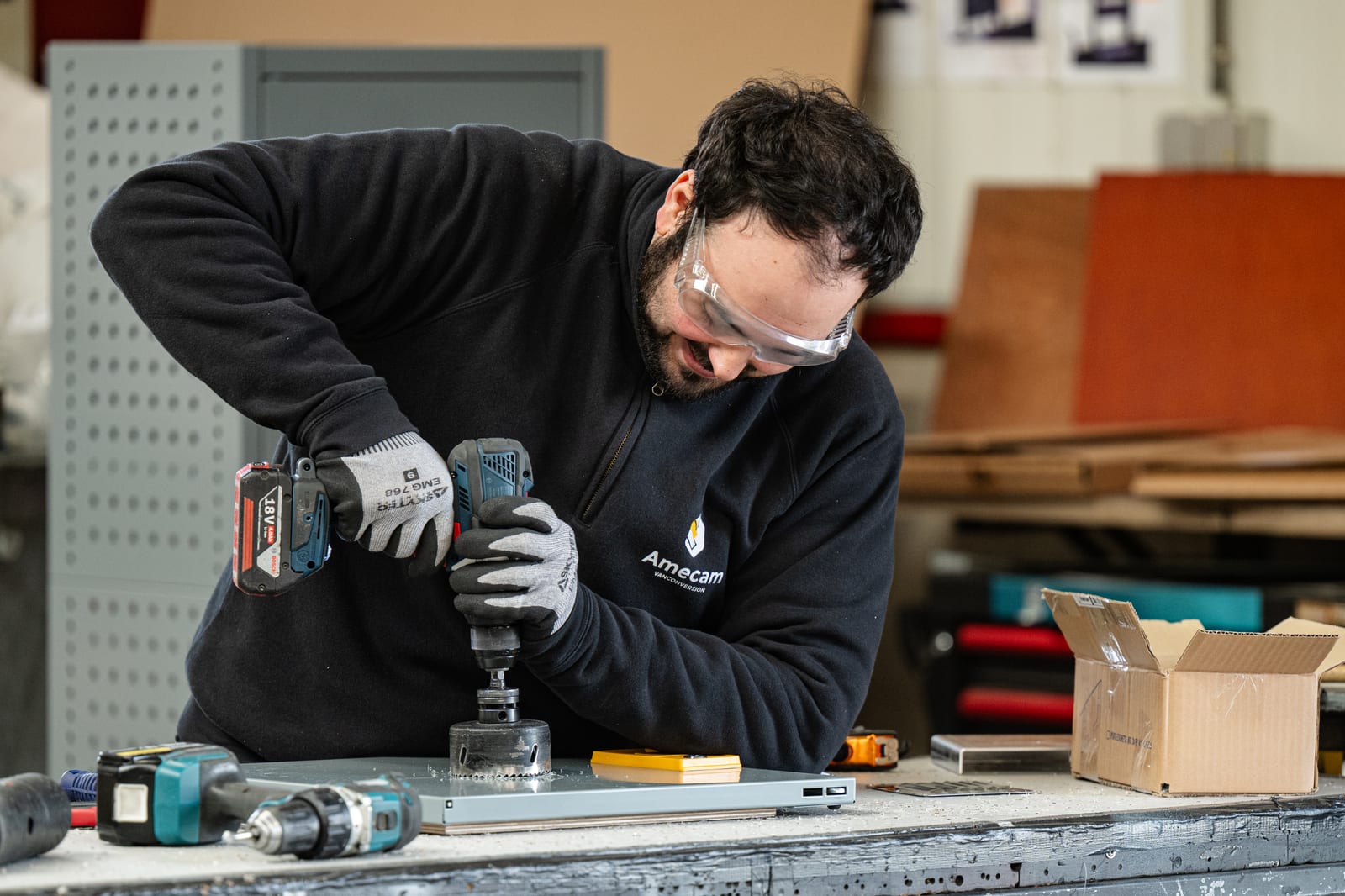 Technicien Amecam perçant une pièce de tôle à la scie-cloche sur l’établi de l’atelier, lunettes de protection